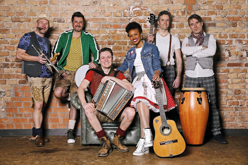 Musikgruppe vor einer Ziegelwand: Links steht ein Mann mit Trompete in der Hand. Er trägt Lederhosen, ein blaues T-Shirt und einen Strohhut und hat eine Zigarre im Mund. Rechts daneben steht ein Mann in Lederhosen, gelbem T-Shirt, grüner Sportjacke, der sich auf einer Trommel abstützt. In der Mitte sitzt ein Mann in Lederhosen und rotem T-Shirt mit einer Diatonischen Harmonika. Neben ihm sitzt eine kubanische Sängerin im Dirndl mit Jeansjacke und einer Gitarre in der Hand. Rechts neben ihr stehen zwei Männer in karierten Hosen, mit hellen Hemden. Vor ihnen steht eine Conga aus Holz.