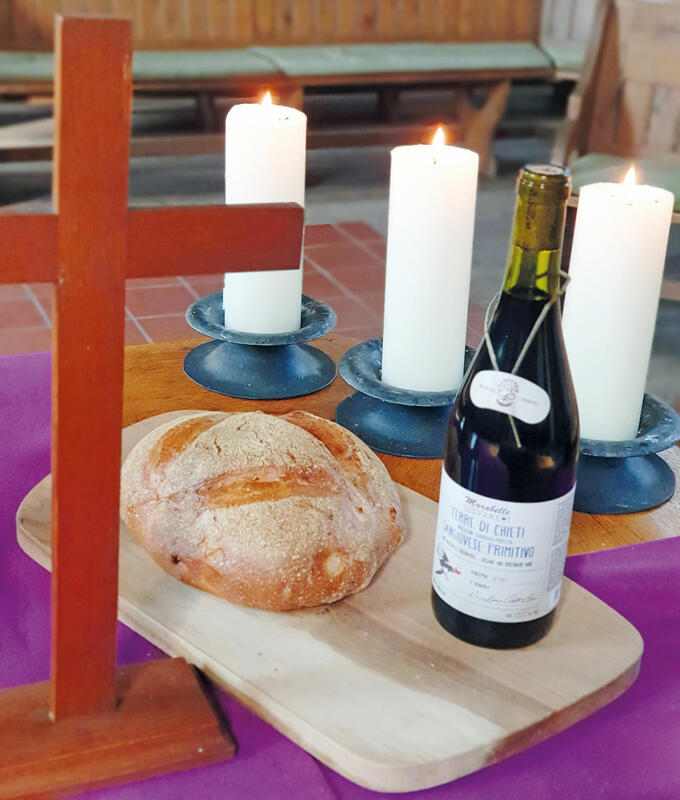 Altartisch aus Holz mit violetter Decke, auf der ein Kreuz, ein Brett mit einer Flasche Wein und einem Brot sowie drei brennende weiße Kerzen stehen.