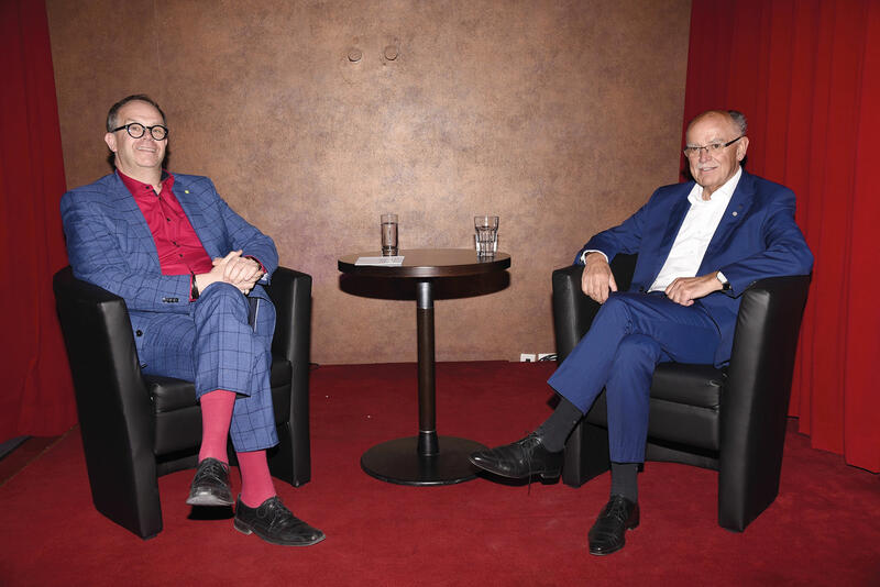 In einem Raum mit rotem Teppichboden sitzen zwei Männer in blauen Anzügen in schwarzen Ledersesseln. Beide tragen Brille. Der linke Mann trägt ein rotes Hemd und rote Socken, der rechte ein weißes Hemd und dunkle Socken. Zwischen ihnen steht ein runder schwarzer Tisch mit zwei Wassergläsern.