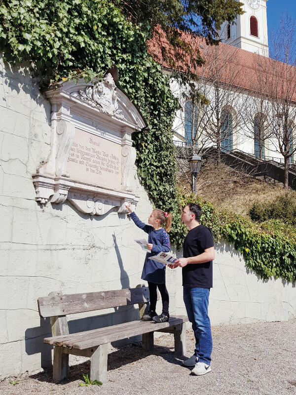 Ein Mann in Jeans und schwarzem T-Shirt betrachtet mit einem Mädchen eine verzierte Inschrift, die an einer Mauer angebracht ist. Das Mädchen steht auf einer Parkbank und trägt ein blaues Kleid, schwarze Strumpfhosen und einen Pferdeschwanz. Über dem Denkmal rankt Efeu. Im Hintergrund ist eine Kirche zu sehen.
