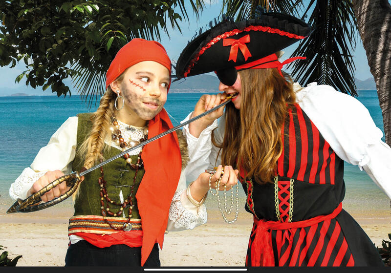 Das Bild zeigt zwei Kinder in weißen Hemden und schwarz-roten Oberteilen vor Meeresstrand und Palmen. Das linke Kind trägt ein rotes Tuch um den Kopf und hat einen Degen in der Hand, das rechte trägt einen schwarz-roten Piratenhut und eine schwarze Augenklappe.