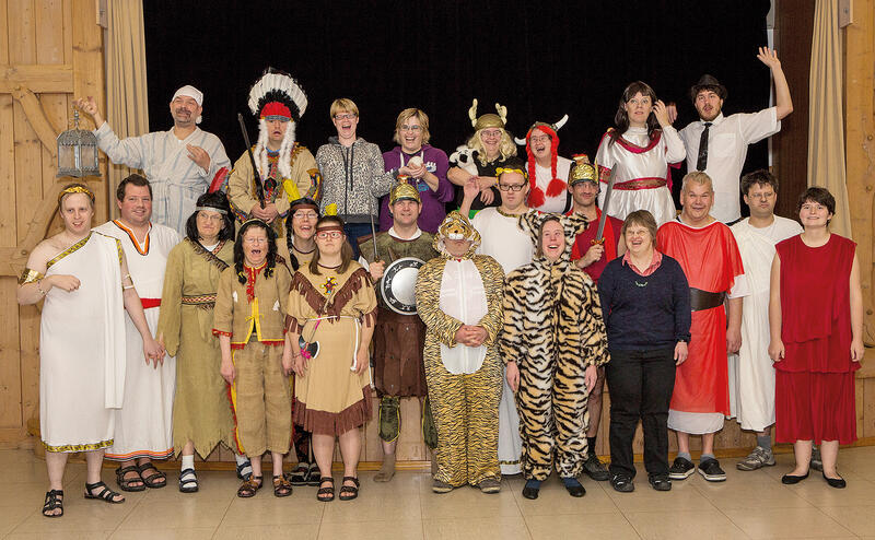 Bühnenbild mit einer in zwei Reihen aufgestellten Theatergruppe, darunter Personen in weißen, beigen und roten Kleidern, ein Indianer und zwei Personen in Tierkostümen