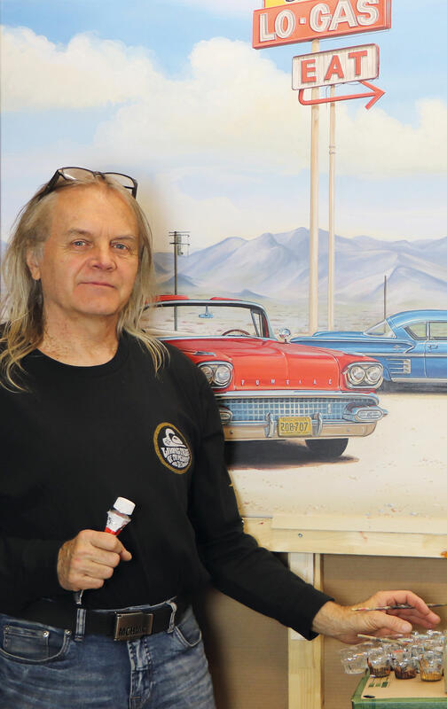 Mann mit langen grauen Haaren, schwarzem Pullover und Jeans hält einen Pinsel in der linken und eine Farbtube in der rechten Hand. Im Hintergrund ein Gemälde mit einem roten und einem blauen amerikanischen Straßenkreuzer vor Bergen