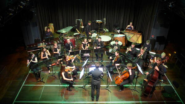 Von erhöhter Position aus sieht man ein Orchester mit Streichern, Bläsern, Schlagwerk und einem Dirigenten
