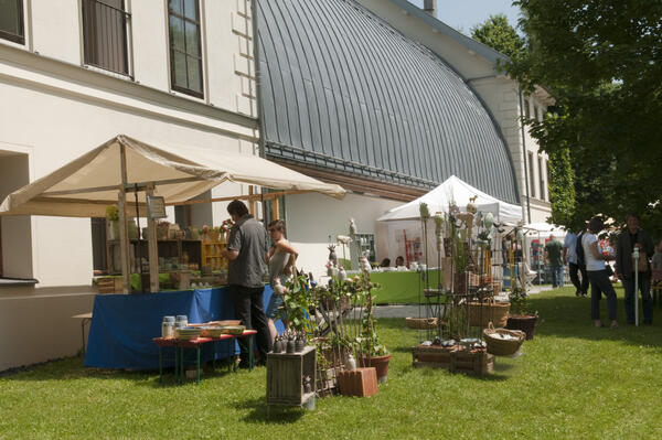 Vor einem Gebäude mit einem markanten tonnenförmigen Dach sind Marktstände aufgebaut, an denen Kunsthandwerksprodukte angeboten werden.