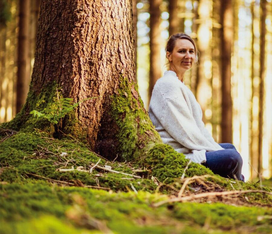 Eine Frau sitzt im Wald an einem Baum. Sie trägt eine weiße Jacke und eine blaue  Jeans. Sie sitzt im Schneidersitz hinter ihr ist ein Baum. Sie lehnt sich dort ab. Im Hintergrund sind Bäume und die Sonne scheint durch. 