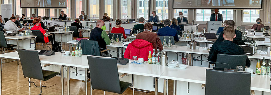 Blick in einen modernen Sitzungssal mit hohen Fenstern:
Viele Personen an Tischen sind von hinten zu sehen. Sie tragen Masken. An der Rückwand sind große Videoleinwände zu sehen.