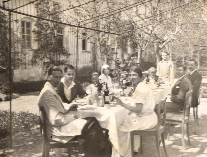 Leicht unscharfes historisches Schwarz-Weiß-Foto von einer Essensgesellschaft im Freien. Männer und Frauen an einem Tisc mit weißer Tischdecke unter einerm Metallgestänge. Im hitergrund Bäume und ein Haus.