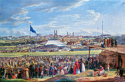 Das historische Bild zeigt eine Ansicht des Oktoberfestes von oben. Im Hintergrund des Bildes sind die Wahrzeichen Münchens zu sehen, darüber ein blauer leicht bewölkter Himmel. Die Personen tragen sehr bunte Klamotten. 