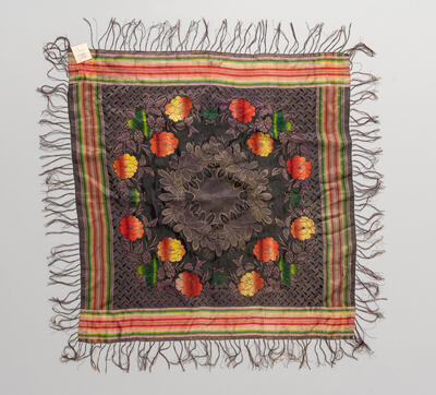 Foto eines quadratischen Tuches in braun mit bunten Streifen an den Kanten und rot gelben Blumenmuster. 