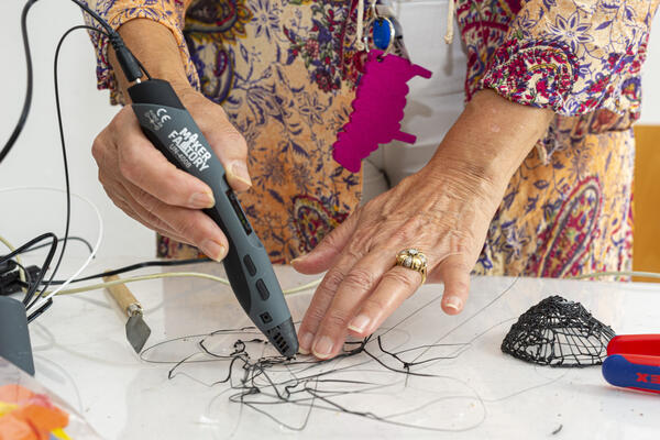 Eine Frau hält in einer Hand einen Stift, aus dem ein schwarzer Kunststofffaden kommt. Mit der anderen Hand modelliert sie ihn zu einem kunstvollen Netz.