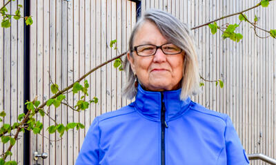 Ältere Frau mit Brille in einem blauen Trainingsanzug im Freien vor einer Wand aus Holzverkleidung