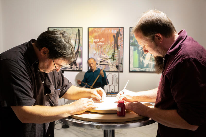 Zwei Männer sitzen an einem runden tisch und zeichnen auf ein weißes, rundes Blatt Papier. Im Vordergrund steht ein kleines Glas rote Farbe. Im Hintergrund sieht man einen Mann der auf einem Stuhl sitzt und Musik macht. Hinter ihm hängen 3 bunte Bilder an der Wand.