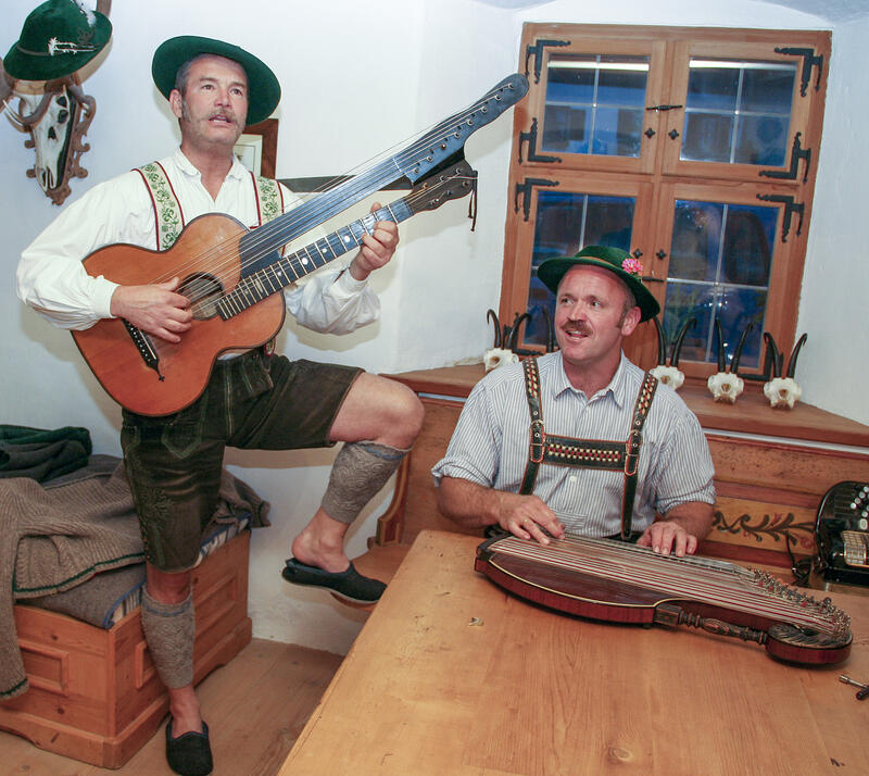 Zwei Musiker in Lederhosen und mit Hut spielen in einer Stube Gitarre und Zither