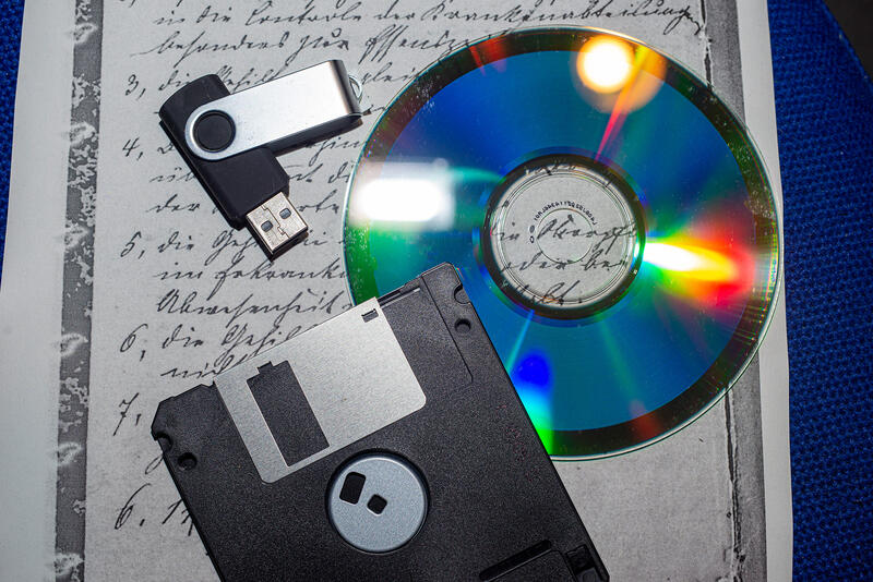 Auf einem Schriftstück, bei dem in altdeutscher Handschrift eine Liste zu sehen ist, liegt eine 3,5"-Diskette, eine DVD und ein USB-Stick.