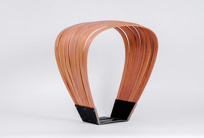 Gebogene Holzstreifen formen einen Bogen, der unten von einem eckigen Metall gefasst wird. Oben ist der Holzbogen flacher und bildet eine Sitzfläche aus Einzelstreifen.  