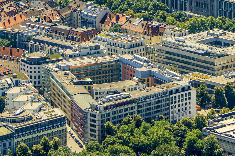 Blick von schräg oben auf München: Im Vordergrund hinter Bäumen ein Block mit Bürogebäuden. Im Hintergrund historische Häuser