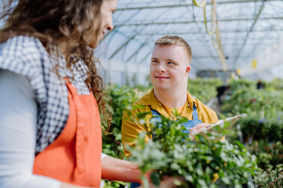 Junger Mann im gelben Shirt steht in einer Gärtnerei. Im Hintergrund sind grüne Pflanzen zu sehen. Im Vordergrund sieht man seine Ansprechpartnerin von der Seite, die den jungen Mann anlächelt.