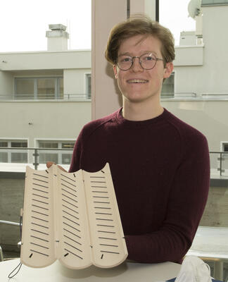 Junger Mann mit Brille und rotem Pullover hält einen zylindrischen Gegenstand aus Holz mit Gummibändern in die Kamera. Im Hintergrund sieht man den Balkon eines Bürogebäudes.