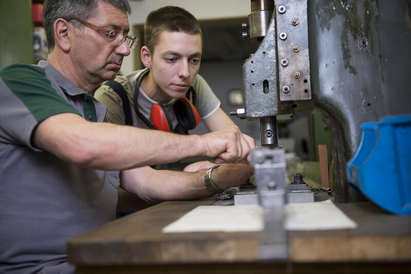 Ein älterer zeigt einen jüngeren Mann etwas an einer Maschine zur Metallverarbeitung.