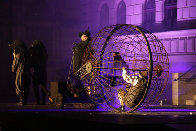 Szene auf der Bühne: Eine kugelförmiger Käfig, in dem ein Mann sitzt. Davor eine Frau in schwarzem Mantel, davor Schauspieler mit Pferdeköpfen an der Deichsel. 