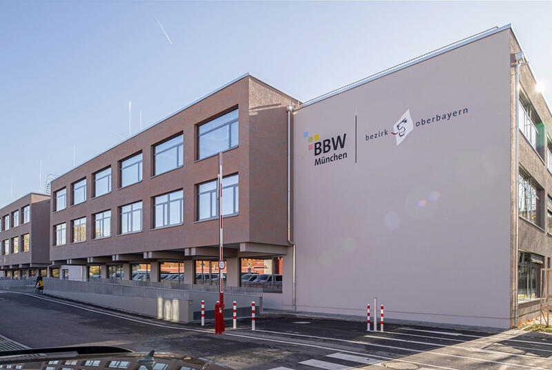 Ein Gebäudekomplex aus mehreren Baukörpern und großen Fensterflächen trägt auf einer Fassadenfläche das Logo des Bezirks Oberbayern neben dem Logo des BBW München