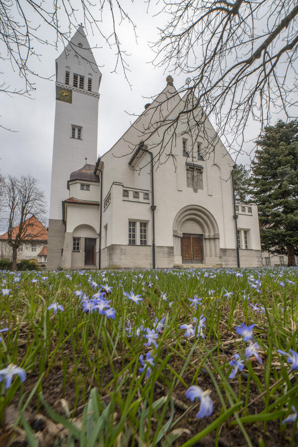 Außenaufnahme einer Kirche mit Kirchturm; im Vordergrund wachsen blaue Blumen im Gras
