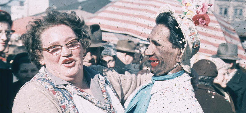 Historisches Foto in verblichenen Farben aus den fünfziger Jahren: Eine Frau und ein Mann umarmen sich auf einem Marktplatz. Die Frau schaut dabei in die Kamera. Der Mann schaut auf die Frau. Er ist offensichtlich in Faschingsverkleidung als Frau.