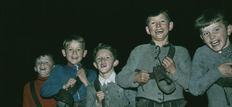 Historisches Foto in verblichenen Farben aus den fünfziger Jahren: Fünf Kinder schauen in die Kamera. Alle haben Kuhglocken um den Hals hängen. Der Hintergrund ist schwarz.