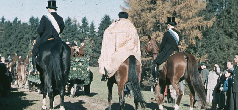 Historisches Foto in verblichenen Farben aus den fünfziger Jahren: Es ist ein Pferdeumzug zusehen. Aber man kann die Pferde und Menschen nur von hinten sehen. Am Rand stehen Menschen, die dem Umzug zuschauen. Im Hintergrund sind Bäume und Wiese zu sehen.