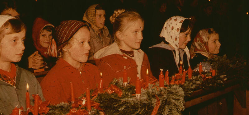 Historisches Foto in verblichenen Farben aus den fünfziger Jahren: Kinder stehen in Kirchbänken. In der ersten Reihe liegen auf der Ablage Kränze aus Tannenzeigen und mit jeweils vier roten Kerzen drauf. Jeweils eine der Kerzen brennt. Die Meisten der Kinder haben ein Kopftuch auf!