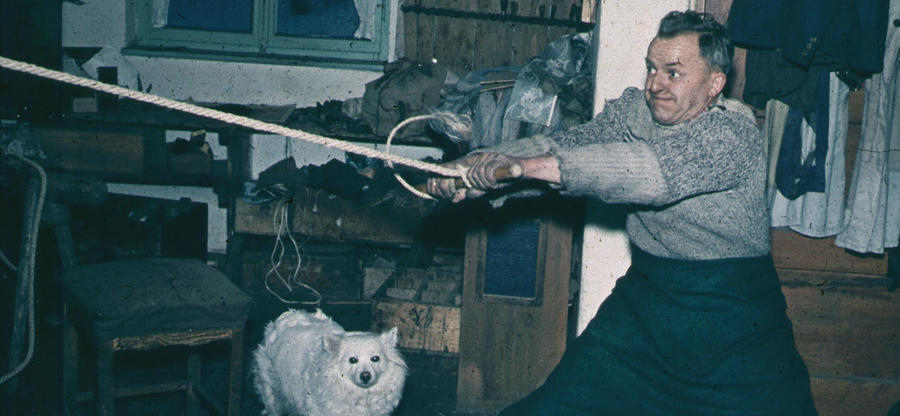 Historisches Foto in verblichenen Farben aus den fünfziger Jahren: Ein Mann ist in einer Werkstatt. Er zieht an einem Stück Holz, das an einem Seil befestigt ist. Links von ihm ist ein kleiner, weißer Hund.