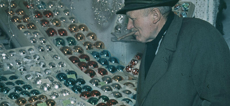 Historisches Foto in verblichenen Farben aus den fünfziger Jahren: Ein Mann mit Zigarre in Mund schaut sich an einem Stand Weihnachtsbaumkugeln an. Die Kugeln haben die Farbe rot, silber, blau und gold.