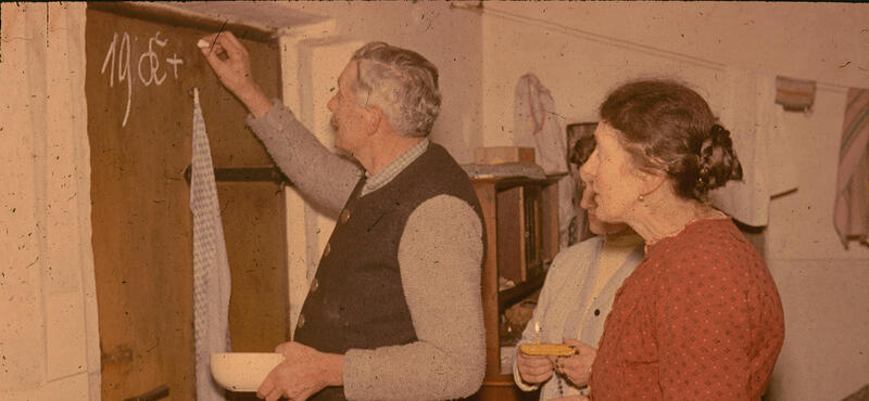 Historisches Foto in verblichenen Farben aus den fünfziger Jahren: Ein Mann schreibt mit Kreide traditionelle Buchstaben an eine Holztüre. Zwei Frauen stehen hinter ihm. Eine von Ihnen hat etwas in der Hand. Unter anderem eine Gebetskette.