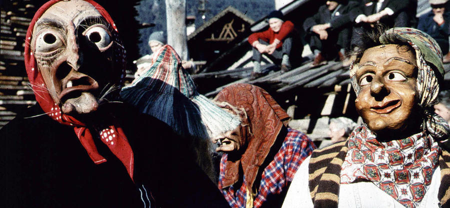 Historisches Foto in verblichenen Farben aus den fünfziger Jahren: Zwei Personen schauen in die Kamera. Ihre Gesichter sind nicht zu erkennen, da sie Masken tragen und verkleidet sind. Im Hintergrund sind weiter Personen, die verleidet sind und eine Maske tragen.