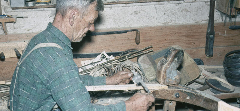 Historisches Foto in verblichenen Farben aus den fünfziger Jahren: Ein Mann arbeitet mit seinen Händen und einem Werkzeug an einem Stück Holz. Im Hintergrund sind noch andere Werkzeuge zu sehen.
