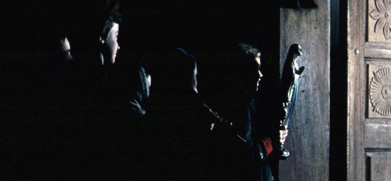 Historisches Foto in verblichenen Farben aus den fünfziger Jahren: Kinderstarr stehen im Dunkeln. Sie sind kaum zu erkennen. Durch eine Holztür kommt etwas Licht herein und lässt von wenigen das Profil des Gesichts erkennen. Das Kind, das am nächsten an der Tür steht hat eine Figur in der Hand.