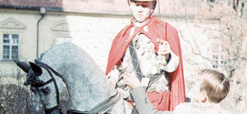Historisches Foto in verblichenen Farben aus den fünfziger Jahren: Ein Mann in Ritterrüstung sitzt auf einem Pferd. Er trägt außerdem einen roten Umhang. Und in einer Hand hält er ein Schwert nach oben.