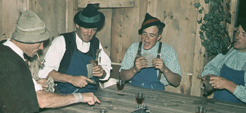Historisches Foto in verblichenen Farben aus den fünfziger Jahren: Vier Männer sitzen an einem Tisch und spielen Karten. Auf dem Tisch stehen außerdem kleine Gläser mit Schnaps. Zwei der Männer haben eine Pfeife im Mund. Alle haben die gleiche Art von Hut auf dem Kopf.