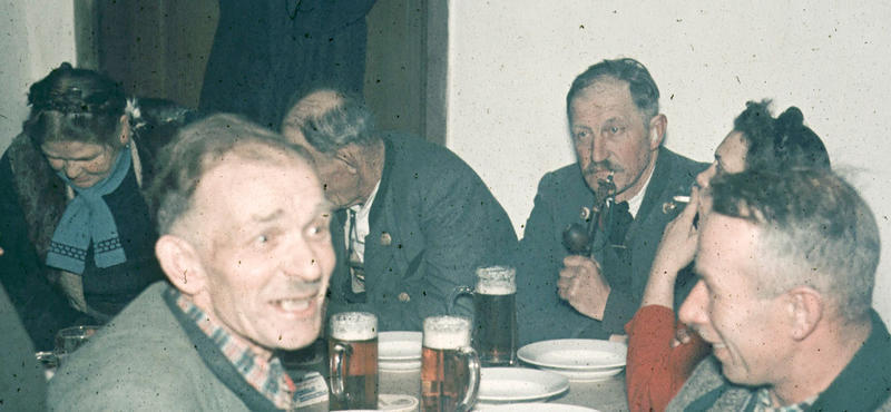 Historisches Foto in verblichenen Farben aus den fünfziger Jahren: Personen sitzen am Tisch. Einer raucht Pfeife und eine andere eine Zigarette. Auf dem Tisch stehen Gläser mit Bier und Suppenteller.