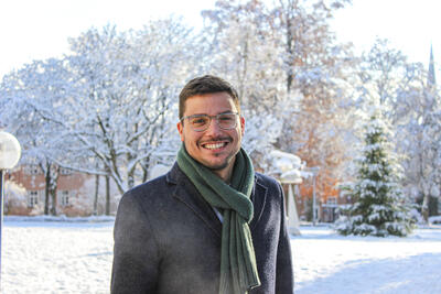 Foto eines jungen, freundlich lachenden Mannes mit Brille. Im Hintergrund ist eine Schneelandschaft zu erkennen. 