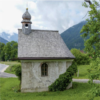 Kleine Kapelle mit Glockentürmchen, halbrunder Apsis und Schindeldach. Die Kapelle steht auf einer Wiese am Wegrand, im Hintergrund sind Berge zu erkennen. Das Mauerwerk wird teils umrankt.