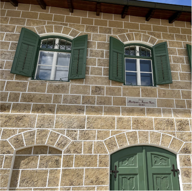 Fassade aus Sandsteinquadern ud aufgemalten weißen Fugen. Ein Ausschnitt der Fassade zeigt zwei Fenster mit grünen Fensterläden, darunter ein Schild mit der Aufschrift "Erbaut Mathias und Anna Latz 1848".