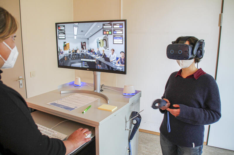 Eine Frau hat eine große VR-Brille auf und einen Joystick in der Hand. Auf einem Monitor ist ein Seminarraum mit vielen Personen zu sehen. Monitor und Frau werden von einer weiteren Person, die eine FFP2-Maske trägt, überwacht.