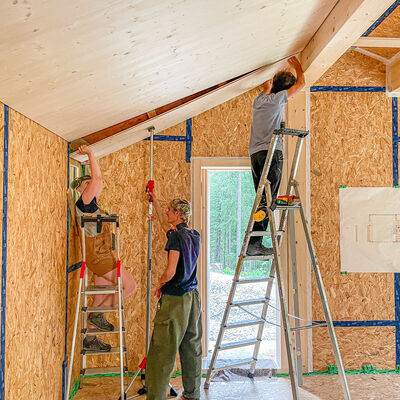 Auf eienr Baustelle: zwei Schreiner und eine Schreinerin besfestigen sie eine Deckenblatte aus Holz in einer Hütte.