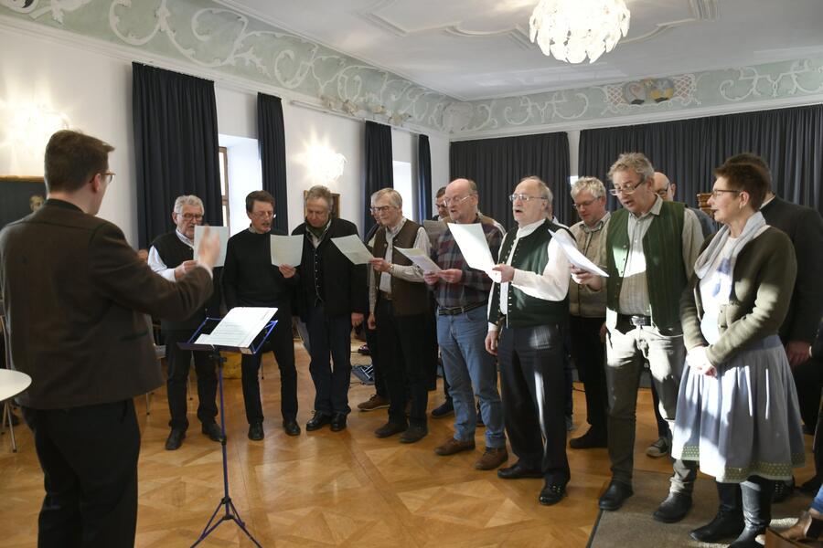 Eine Gruppe von ca. zehn Männern und Frauen stehend im Halbkreis, mit Notenblättern in der Hand; singend; Der Volksmusikpfleger Leonhard Meixner dirigiert. In einem größeren Saal mit Kronleuchter;