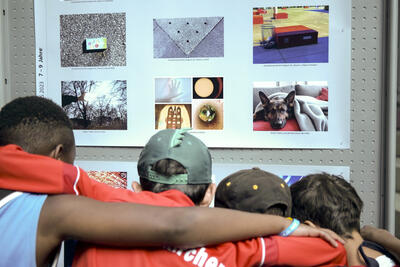 Man sieht vier Kinder von hinten, die mit eingehakten Armen über den Schultern vor einer Ausstellungswand stehen, auf der Fotografien ausgestellt sind.