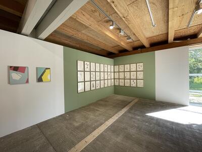 Blick in einen Ausstellungsraum mit Holözecke und grüner Wand mit quadratischen Formaten