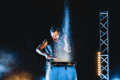 Mann steht vor einer blauen Trommel aus der eien Wasserfontäne hochspritzt währen er mit den Schalgzeugstöcken auf die Trommel einschlägt. das ganze ist auf einer Bühne, die Beleuchtung von schräg unten.