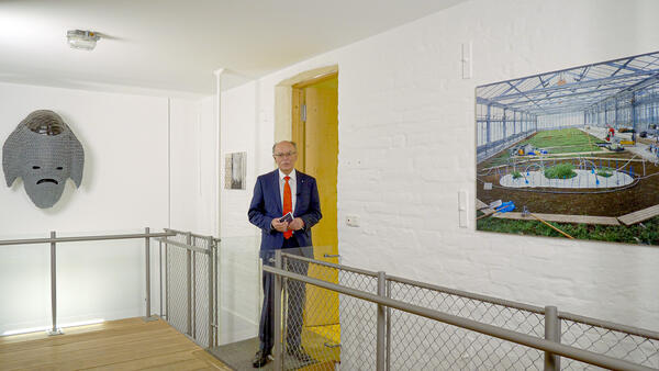 Ein Mann in Anzug spricht zu den Zusehenden. Links und rechts von ihm Sind Kunstwerke an den Wänden zu sehen, die im Rahmen eines früheren 